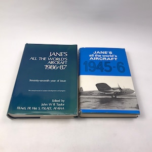 Puede incluir: Dos libros antiguos titulados "Jane's All the World's Aircraft". El libro de la izquierda es azul oscuro y dice "1986-87". El libro de la derecha es azul y dice "1945-6". Ambos libros son de tapa dura y presentan texto e imágenes.