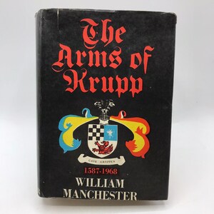 May include: Black book cover with the title "The Arms of Krupp" in white lettering. The cover also features a colorful coat of arms with the text "Cave Gryppen 1587-1968" and the author's name "William Manchester" in white lettering.