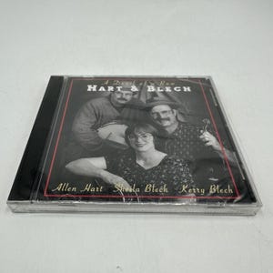 May include: A CD case featuring the album "A Devil of a Row" by Hart & Blech. The cover shows three people, with the band's name and member names Allen Hart, Sheila Blech, and Kerry Blech printed on the cover.
