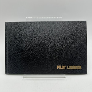 May include: Black Pilot Logbook with gold lettering. The logbook has a textured cover and is rectangular in shape. The text "PILOT LOGBOOK" is printed in gold on the front cover.