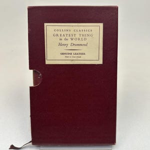 May include: A burgundy-colored book in a matching slipcase. The book's title, "GREATEST THING in the WORLD" by Henry Drummond, is displayed on a cream-colored label. The label also states "GENUINE LEATHER" and "Made in Great Britain."