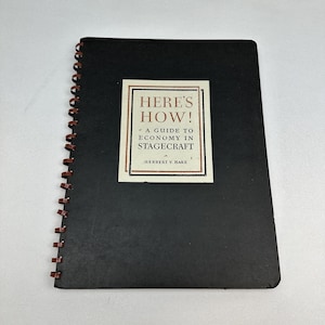 Op de afbeelding: Een zwart spiraalgebonden notitieboek met een rechthoekig etiket op de voorkant. Het etiket luidt "HERE'S HOW! A GUIDE TO ECONOMY IN STAGECRAFT" in bruine tekst. De spiraalbinding is bruin.