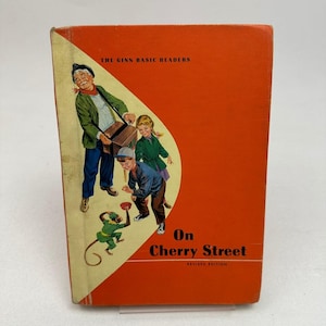 Könnte beinhalten: Vintage Kinderbuch mit dem Titel "On Cherry Street" mit einem leuchtend orangefarbenen Einband. Das Cover zeigt Illustrationen eines Mannes, zweier Kinder und eines Affen. Der Text "The Ginn Basic Readers" steht oben.