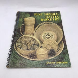 Puede incluir: Un libro encuadernado en espiral titulado "Pine Needle Raffia Basketry" con imágenes de cestas tejidas, una taza y una bandeja con piñas. La portada tiene un fondo verde y el nombre de la autora, Jeannie McFarland.