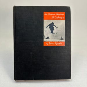 May include: A hardcover book with a black cover. The book has a rectangular orange sticker with the title "The Hannes Schneider Ski Technique" and an image of a skier. The author's name, Benno Rybicka, is also printed on the sticker.
