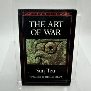May include: A black hardcover book with the title "THE ART OF WAR" by Sun Tzu. The cover features a green and brown graphic and the text "Shambhala Pocket Classics". The book is translated by Thomas Cleary.