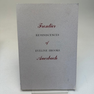 May include: A light gray book cover featuring the title Frontier REMINISCENCES of EVELINE BROOKS Auerbach printed in a combination of dark red script and black serif capital letters centered on the front surface of the rectangular book.