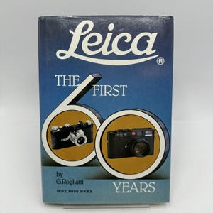Puede incluir: Un libro titulado "Leica The First 60 Years" de G.Rogliatti. La portada presenta el logotipo de Leica en blanco, un fondo azul y dorado, e imágenes de dos cámaras Leica negras. También se ve el texto "HOVE FOTO BOOKS".
