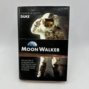May include: A hardcover book titled "Moon Walker" by Charlie & Dotty Duke. The cover features an astronaut saluting, with the Earth and moon in the background. The book's description is about an astronaut's journey.