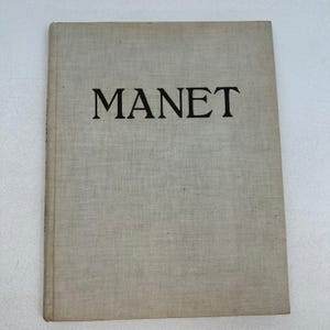 May include: A hardback book with a light beige, textured fabric cover. The word MANET is printed in large, bold, black capital letters across the upper centre of the front cover. The book has a rectangular shape with a visible spine on the left side.