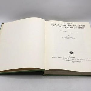 Op de afbeelding: Open boek met de titel "DESIGN AND CONSTRUCTION OF STEEL MERCHANT SHIPS" met groene kaft. De tekst is zwart op gebroken witte pagina's. Uitgegeven door The Society of Naval Architects.