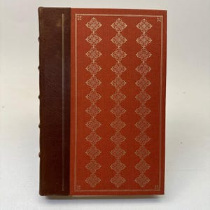 May include: A hardcover book with a red cover featuring a gold geometric pattern. The spine is brown leather. The book is closed and upright, showcasing its decorative design.