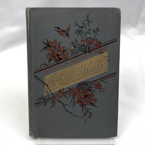 May include: A vintage book cover with a gray background and a gold and red illustration of a bird's nest, flowers, and birds. The title of the book is "Who Was the First Paper-Maker?" and the subtitle is "The Tools and Industries of Animals".