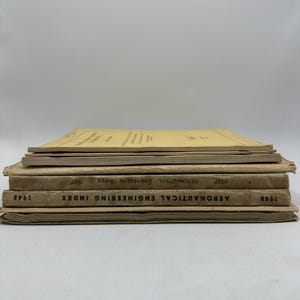 Può includere: Una pila di libri d'epoca sull'indice di ingegneria aeronautica. I libri sono marroni e beige, con testo e date visibili sui dorsi. Il libro in cima è di un beige più chiaro.