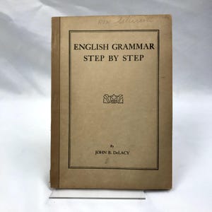 May include: An antique book titled "English Grammar Step by Step" by John B. DeLacy. The book has a tan cover with black text and a decorative emblem. The book's spine is a darker brown.
