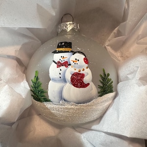 May include: A silver Christmas ornament with a painted scene of two snowmen. The snowman on the left is wearing a black top hat and a red bow tie. The snowman on the right is wearing a red scarf and a red hat. Both snowmen are standing in a snowy scene with green pine trees.
