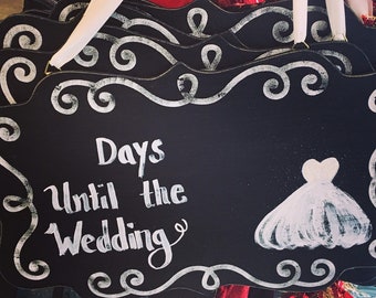May include: Black chalkboard sign with white calligraphy that reads "Days Until the Wedding" and a white silhouette of a wedding dress. The sign is decorated with white swirls.