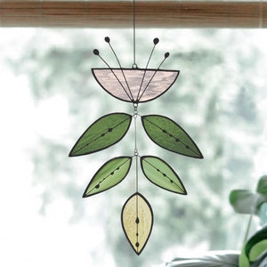 May include: A stained glass window hanging features a pink flower with green leaves. The flower is made of stained glass and has a delicate design. The leaves are also made of stained glass and have a more organic shape.