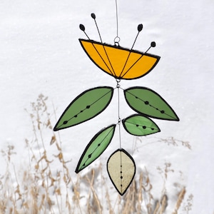 May include: A stained glass mobile featuring a yellow flower-like shape with black accents and green leaf shapes. The leaves and flower have black outlines and are suspended by thin wires. The background is a snowy landscape.