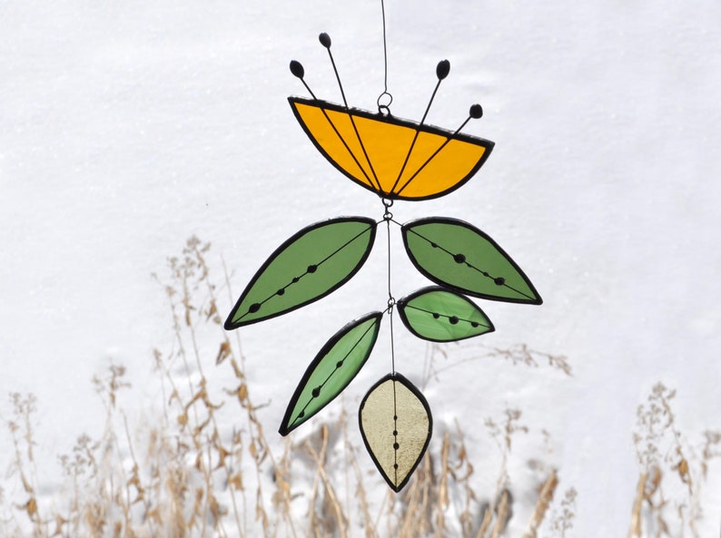 May include: A stained glass mobile featuring a yellow flower-like shape with black accents and green leaf shapes. The leaves and flower have black outlines and are suspended by thin wires. The background is a snowy landscape.
