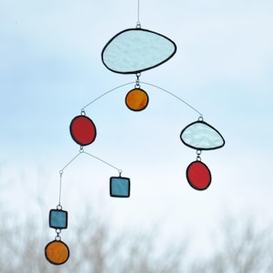 Puede incluir: Un m&oacute;vil hecho de vidrio coloreado con varias formas y colores, incluyendo una nube azul, c&iacute;rculos rojos, c&iacute;rculos naranjas y cuadrados azules.