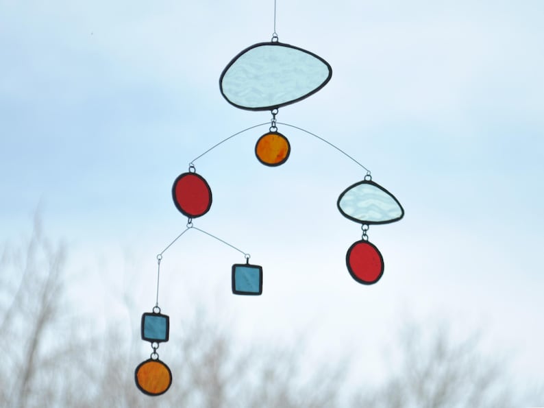 Puede incluir: Un m&oacute;vil hecho de vidrio coloreado con varias formas y colores, incluyendo una nube azul, c&iacute;rculos rojos, c&iacute;rculos naranjas y cuadrados azules.