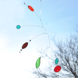 May include: A mobile sculpture with red, green, and brown glass pieces hanging from thin wires against a blue sky background.