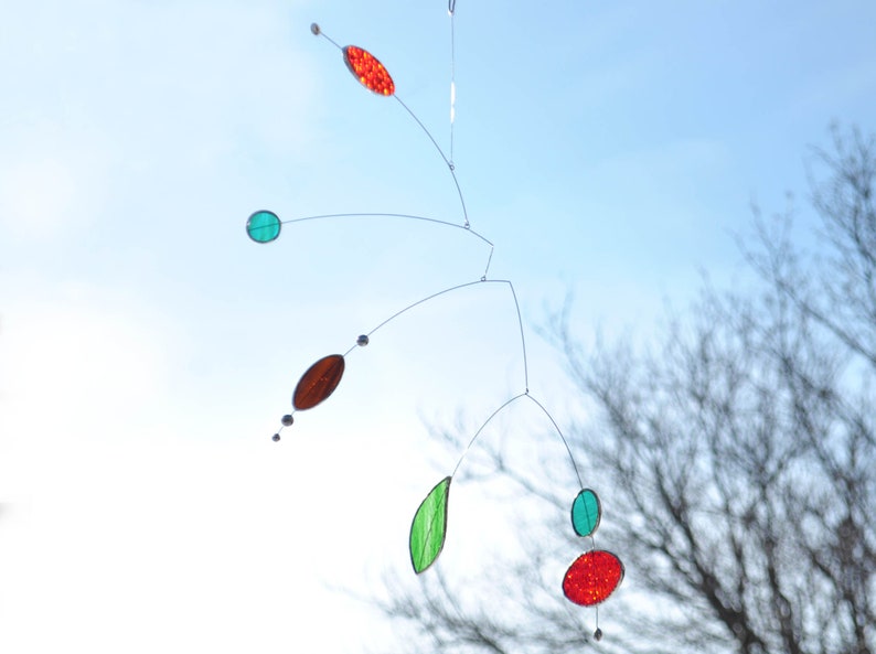 May include: A mobile sculpture with red, green, and brown glass pieces hanging from thin wires against a blue sky background.