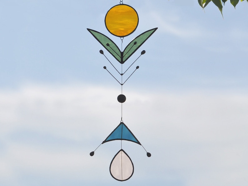Peut inclure: Un mobile en verre coloré avec un cercle jaune, des feuilles vertes, un cercle noir, un triangle bleu et une forme de goutte rose pâle.