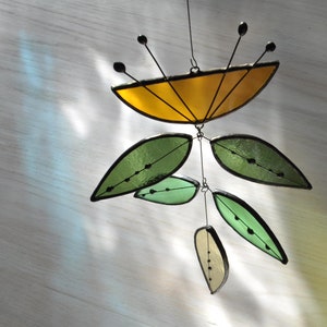 May include: A stained glass mobile with a yellow, semi-circular top and black antennae. Below, green and pale yellow leaf shapes hang from wires. The leaves have black outlines and small black dots. The mobile is designed to catch the light and is suitable for indoor use.
