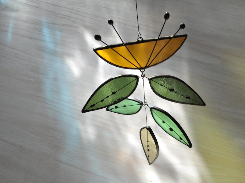 May include: A stained glass mobile with a yellow, semi-circular top and black antennae. Below, green and pale yellow leaf shapes hang from wires. The leaves have black outlines and small black dots. The mobile is designed to catch the light and is suitable for indoor use.