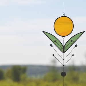 Pode incluir: Um apanhador de sol de vidro colorido com um c&iacute;rculo amarelo e folhas verdes penduradas em uma moldura de metal preto.