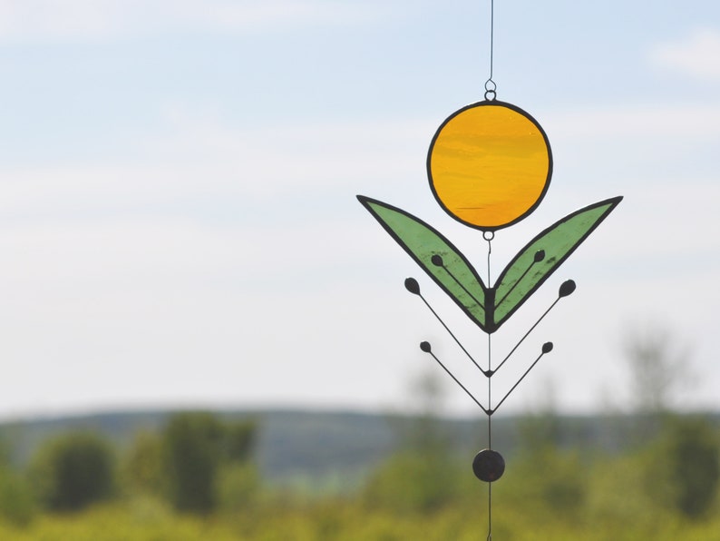 Pode incluir: Um apanhador de sol de vidro colorido com um c&iacute;rculo amarelo e folhas verdes penduradas em uma moldura de metal preto.