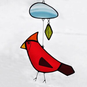 May include: A stained glass cardinal bird with a red body, orange head, yellow beak, and brown tail. The bird is hanging from a blue cloud with a green leaf hanging below.