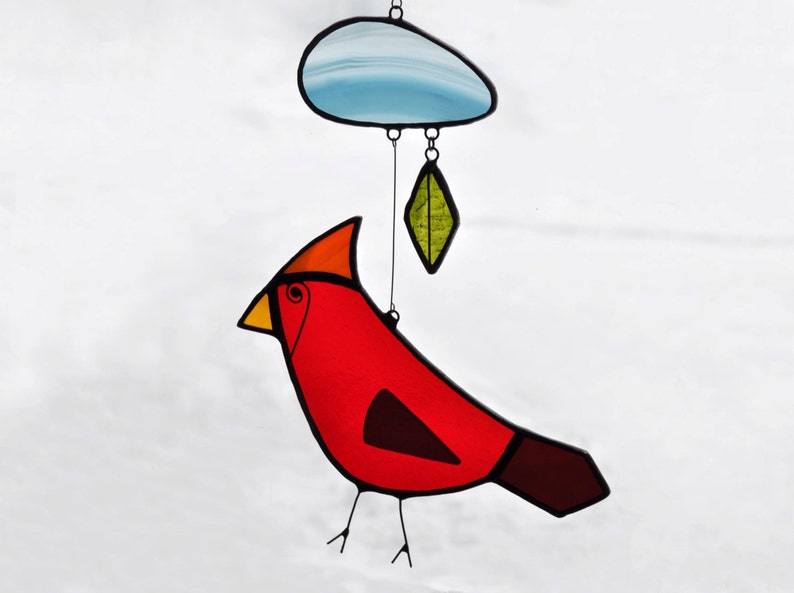 May include: A stained glass cardinal bird with a red body, orange head, yellow beak, and brown tail. The bird is hanging from a blue cloud with a green leaf hanging below.