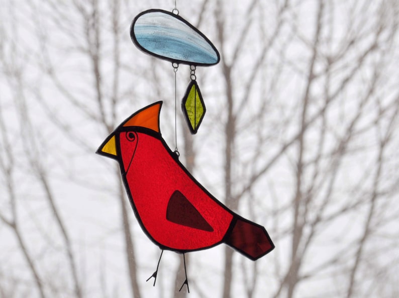 May include: A stained glass cardinal bird mobile with a red body, orange head, and black beak. The bird is hanging from a wire with a blue and white cloud and a green leaf hanging below it.