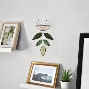 K&ouml;nnte beinhalten: Ein rosa-gr&uuml;nes Glasmobil mit Blumenmuster h&auml;ngt von der Decke. Das Mobile ist an einer wei&szlig;en Wand befestigt, unter der sich zwei wei&szlig;e Regale befinden. Die Regale sind mit gerahmten Bildern und Topfpflanzen dekoriert.
