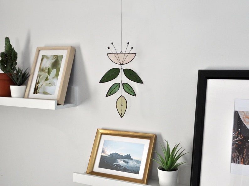 K&ouml;nnte beinhalten: Ein rosa-gr&uuml;nes Glasmobil mit Blumenmuster h&auml;ngt von der Decke. Das Mobile ist an einer wei&szlig;en Wand befestigt, unter der sich zwei wei&szlig;e Regale befinden. Die Regale sind mit gerahmten Bildern und Topfpflanzen dekoriert.