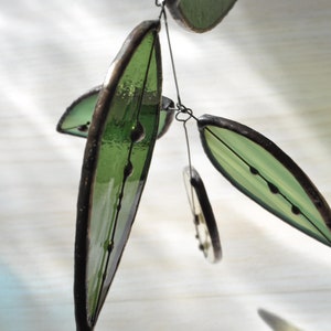 May include: A decorative mobile with green glass leaf shapes and dark brown edging. The leaves are suspended by thin black wire. The glass has a textured finish and small dark details. The background is a light, blurred surface.