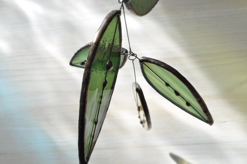 May include: A decorative mobile with green glass leaf shapes and dark brown edging. The leaves are suspended by thin black wire. The glass has a textured finish and small dark details. The background is a light, blurred surface.