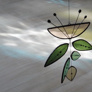 K&ouml;nnte beinhalten: Ein Mobile aus Buntglas mit einem leicht braunen, gebogenen oberen Teil und mehreren gr&uuml;nen Blattformen, die darunter h&auml;ngen. Die Bl&auml;tter haben schwarze Umrisse und kleine schwarze Punkte.