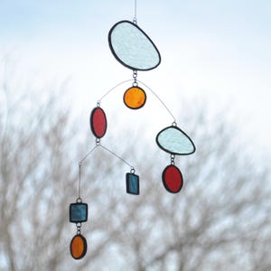 Puede incluir: Un m&oacute;vil hecho de vidrio coloreado con varias formas y colores, incluyendo azul, rojo, naranja y blanco. El m&oacute;vil est&aacute; suspendido del techo y est&aacute; hecho de alambre.
