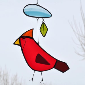 May include: A stained glass cardinal with a red body, yellow beak, and black legs. The cardinal is hanging from a blue cloud with a green leaf hanging below it.