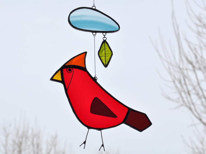 May include: A stained glass cardinal with a red body, yellow beak, and black legs. The cardinal is hanging from a blue cloud with a green leaf hanging below it.