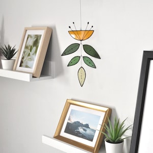 May include: A stained glass mobile with an orange top and green leaf shapes hangs against a white wall. Below, framed pictures and potted plants sit on shelves. The artwork is a decorative piece.