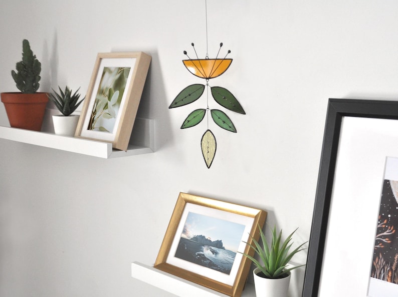 May include: A stained glass mobile with an orange top and green leaf shapes hangs against a white wall. Below, framed pictures and potted plants sit on shelves. The artwork is a decorative piece.