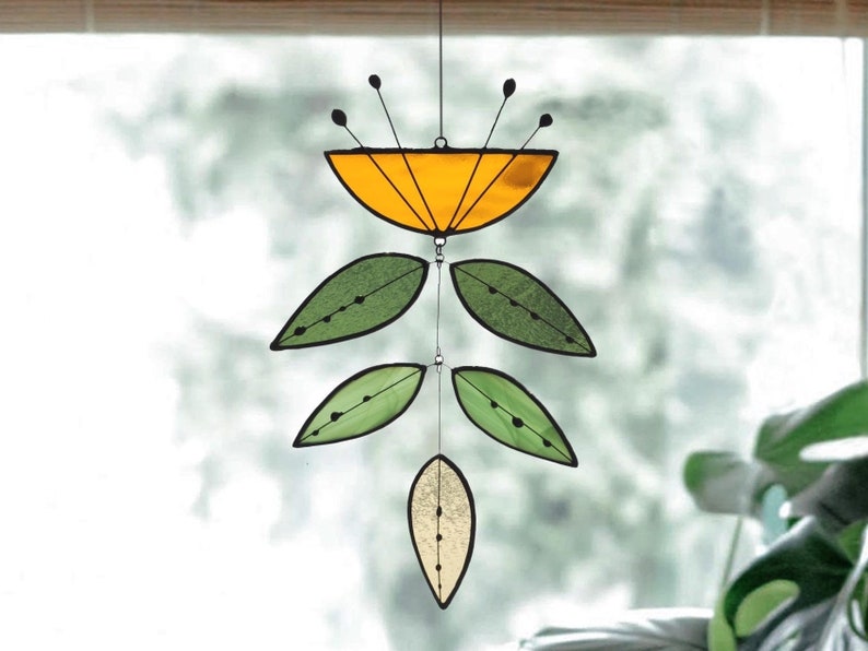 May include: A stained glass suncatcher with a floral design. The top section is a yellow semi-circle with black accents and thin black rods. Below are green leaf shapes and a single, smaller, cream-coloured leaf. The suncatcher hangs from a thin wire.