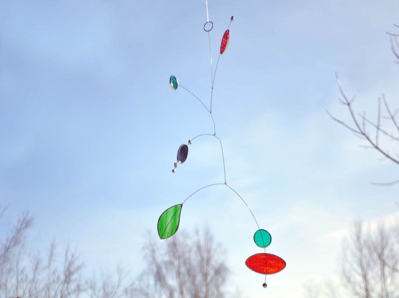May include: A mobile sculpture with a red, green, and purple glass design hanging against a blue sky.