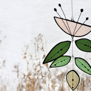 K&ouml;nnte beinhalten: Ein Mobile aus Buntglas mit einer rosa Blume und gr&uuml;nen Bl&auml;ttern. Das Mobile h&auml;ngt an einer schwarzen Schnur vor einem wei&szlig;en Hintergrund.
