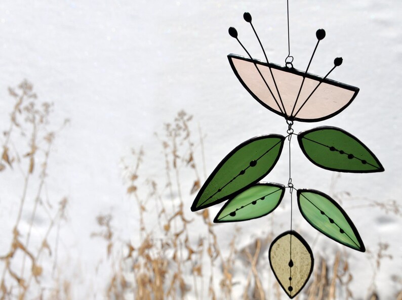 K&ouml;nnte beinhalten: Ein Mobile aus Buntglas mit einer rosa Blume und gr&uuml;nen Bl&auml;ttern. Das Mobile h&auml;ngt an einer schwarzen Schnur vor einem wei&szlig;en Hintergrund.